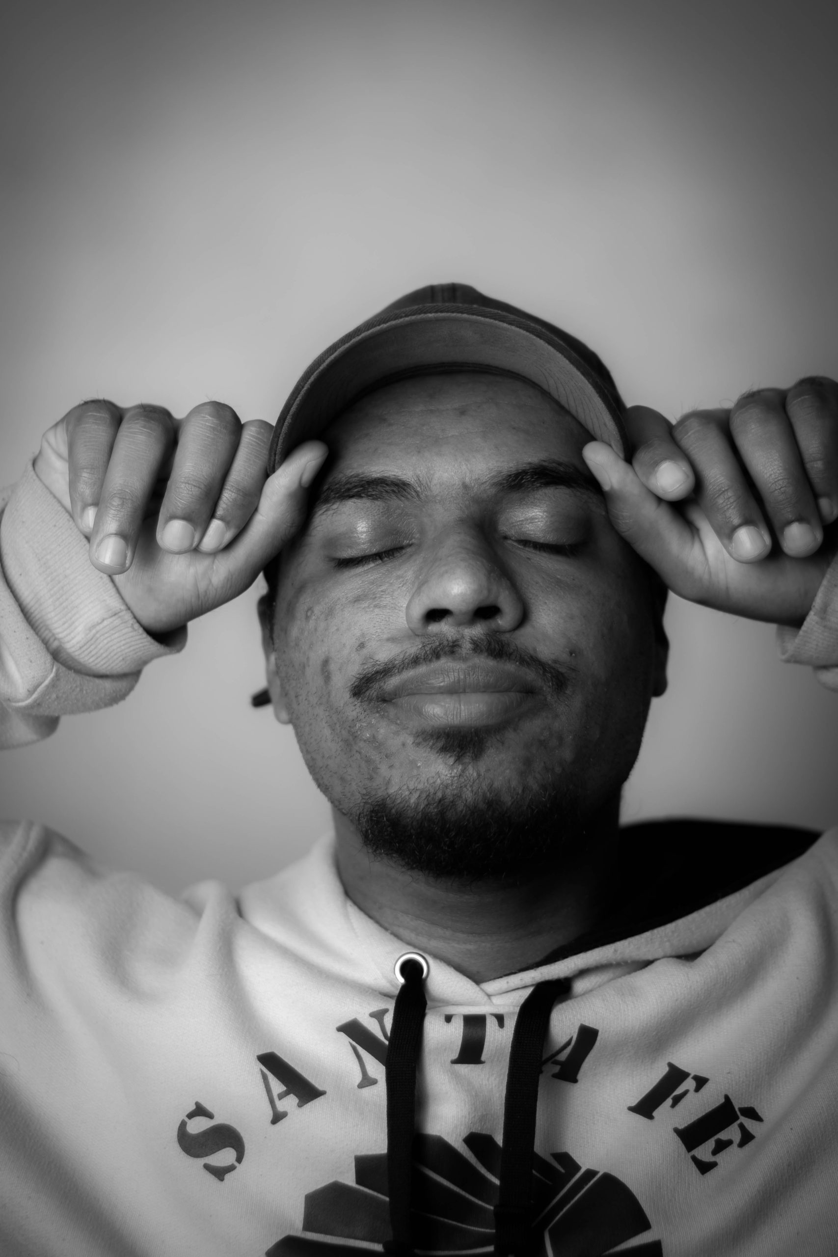 Black and white image of a guy closing his eyes while holding his cap, wearing a sweatshirt that reads